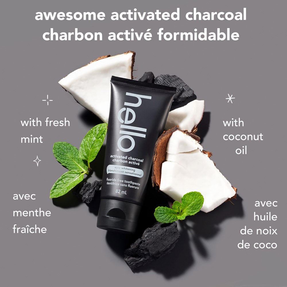 A black tube of hello epic whitening fluoride free charcoal toothpaste is centered, surrounded by pieces of coconut, black charcoal chunks, and fresh mint leaves. The background is light grey. Text above the product reads "awesome activated charcoal." To the left, it says "with fresh mint," and to the right, "with coconut oil."