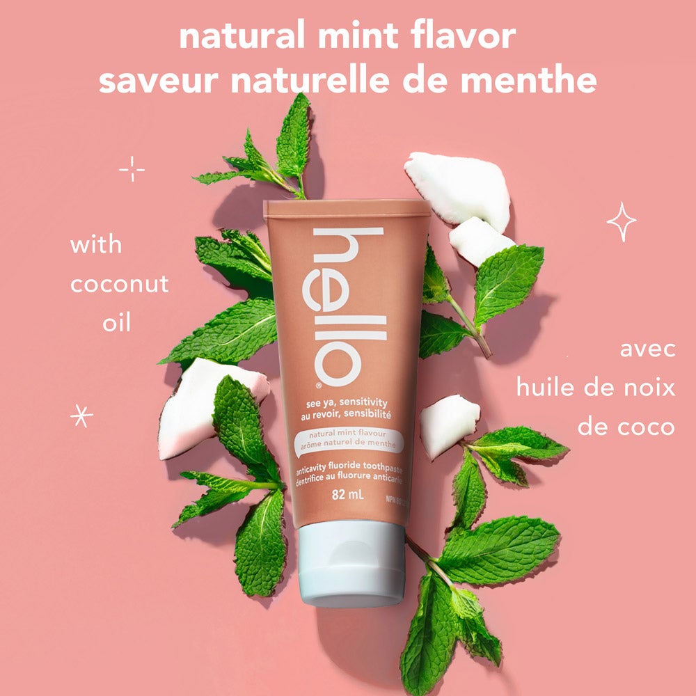 A tube of hello See Ya Sensitivity toothpaste is surrounded by fresh mint leaves and pieces of coconut on a pink background. Text highlights "natural mint flavor" and "with coconut oil" in both English and French.