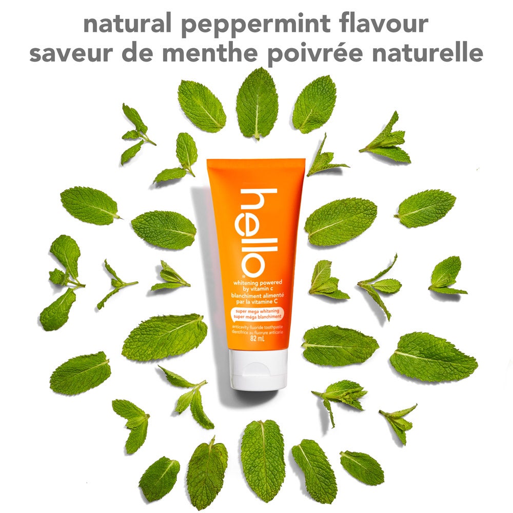 An orange tube of hello super mega whitening toothpaste sits upright in the center, surrounded by fresh peppermint leaves arranged in a circle on a white background. Text at the top says "natural peppermint flavour" in English and French.