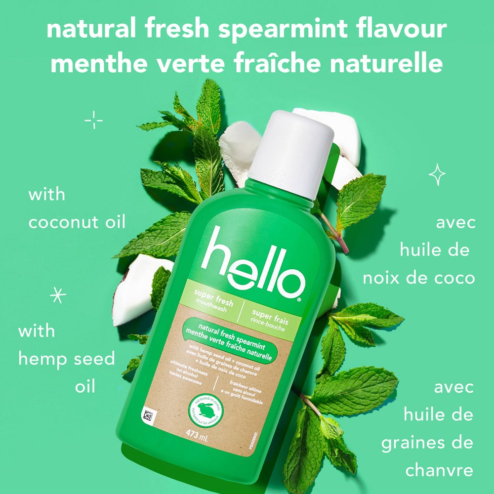 A green bottle of hello super fresh mouthwash is placed on a mint-colored background surrounded by fresh mint leaves, coconut pieces, and hemp seeds. Text says, "natural fresh spearmint flavour" in English and French, with smaller text indicating "with coconut oil" and "with hemp seed oil" in both languages.