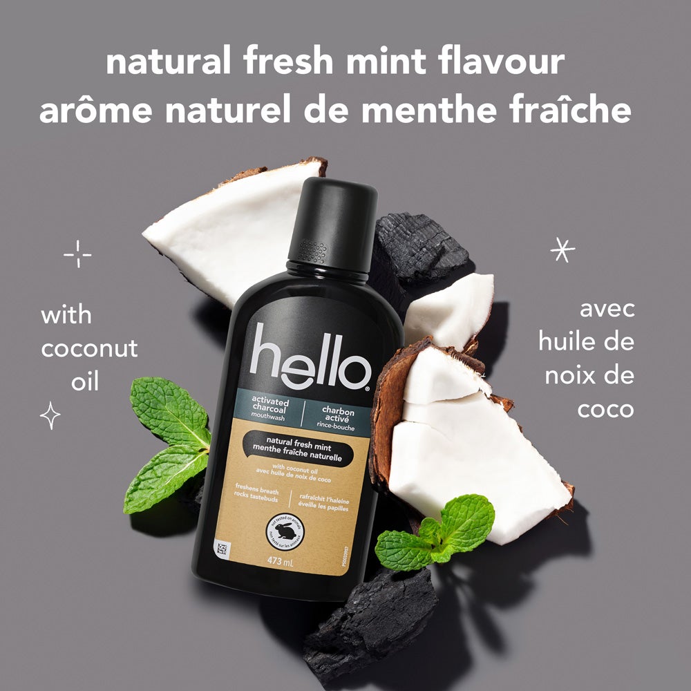 A black bottle of hello activated charcoal mouthwash is centered on a light grey background, surrounded by pieces of coconut, black charcoal chunks, and fresh mint sprigs. The text above reads: "natural fresh mint flavour." To the left, it says "with coconut oil."